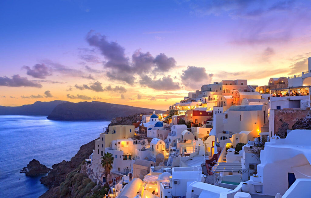 A breathtaking sunset over the Aegean Sea in Santorini, Greece, with the sky painted in vibrant hues of orange, pink, and purple, and the silhouette of whitewashed buildings on the cliffside