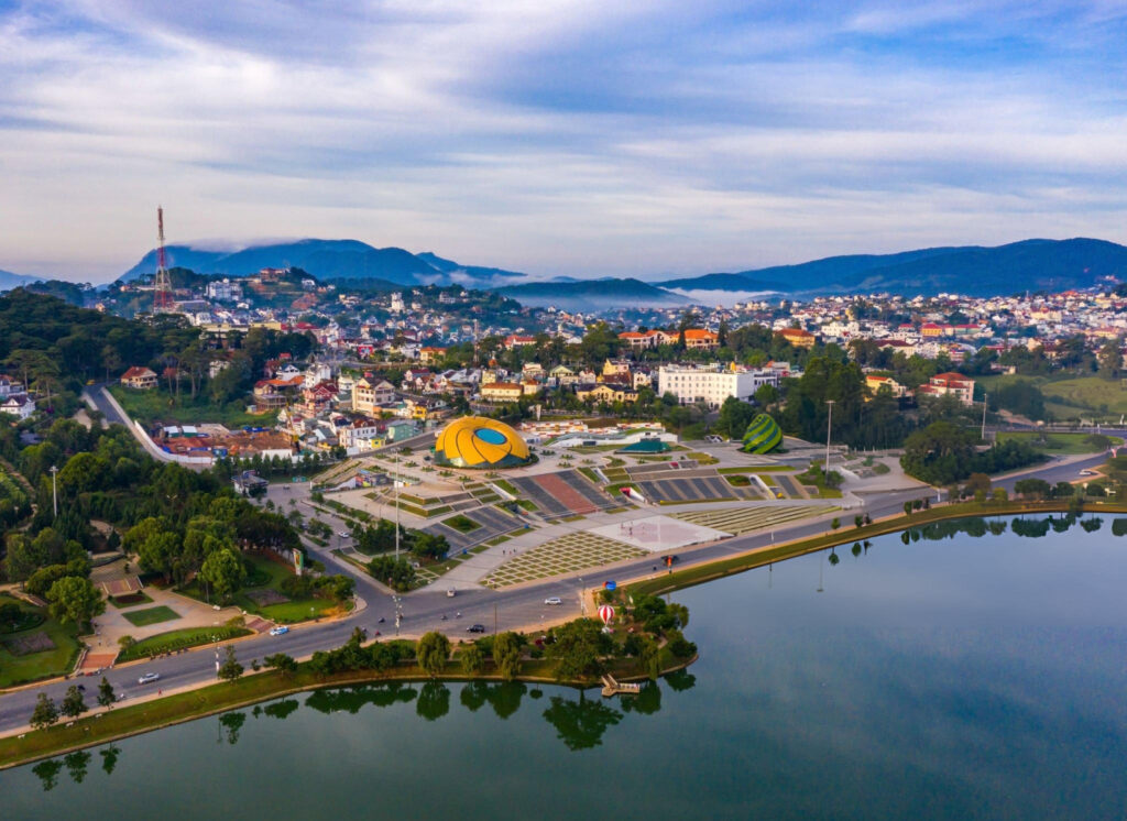 A panoramic view of Dalat, Vietnam, showcasing the city's lush green landscape, hills, and serene lakes with a few colonial-style buildings scattered around