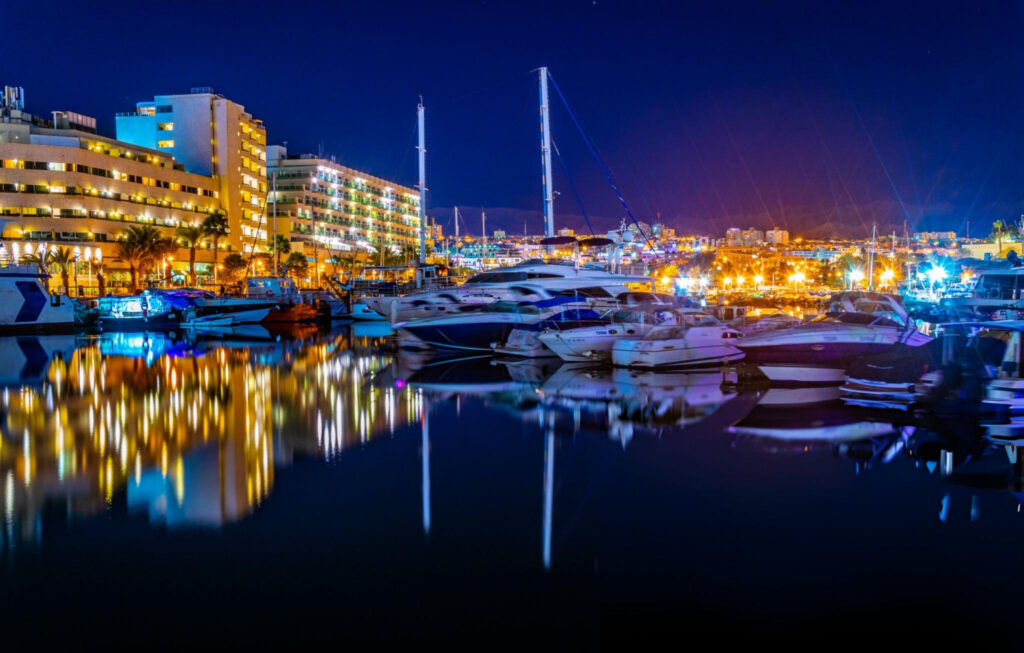 A nighttime view of a luxury resort in Eilat, Israel, featuring a beautifully illuminated swimming pool, ambient lighting around the pool area, and the city lights reflecting on the calm waters of the Red Sea.