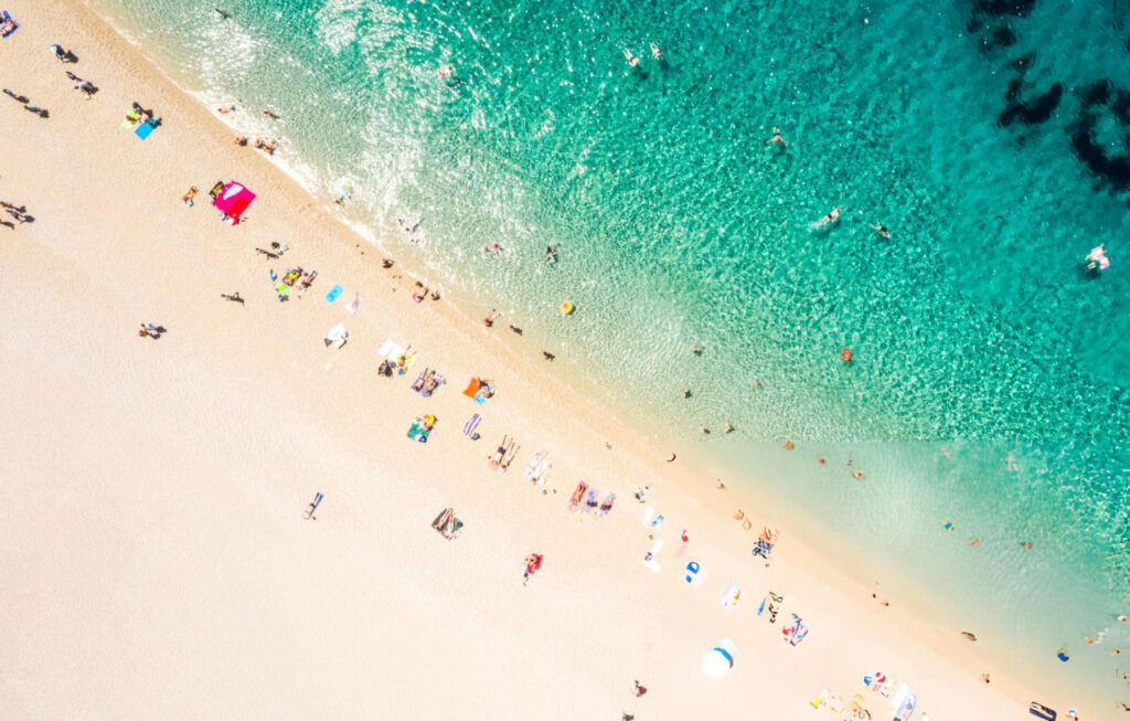 A panoramic view of Zlatni Rat Beach in Bol, Croatia, featuring crystal-clear turquoise waters, golden sand, and lush greenery in the background