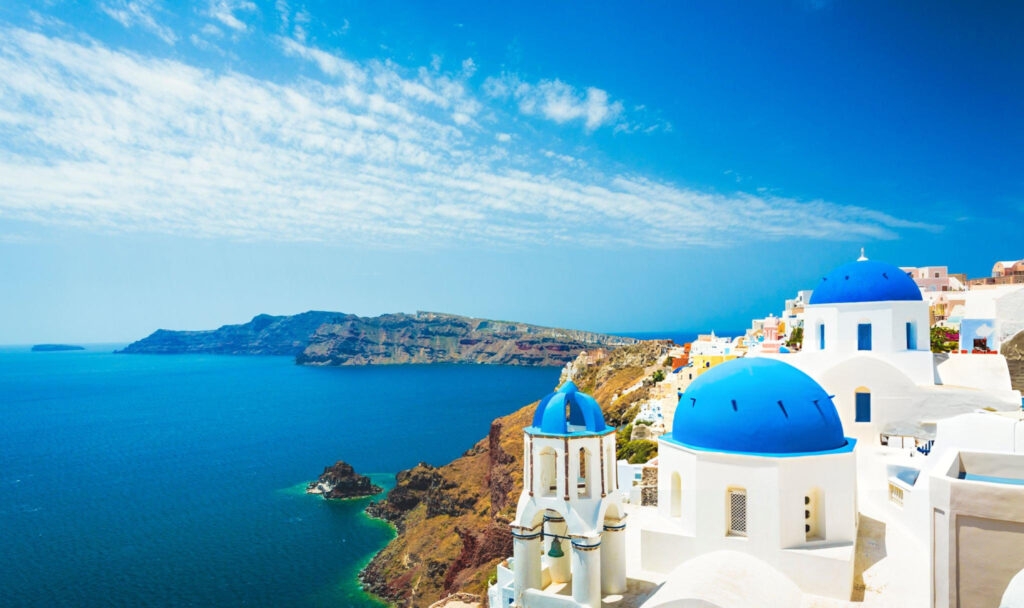 A picturesque view of Santorini Island, Greece, featuring whitewashed buildings with blue domes perched on cliffs overlooking the deep blue Aegean Sea under a clear sky