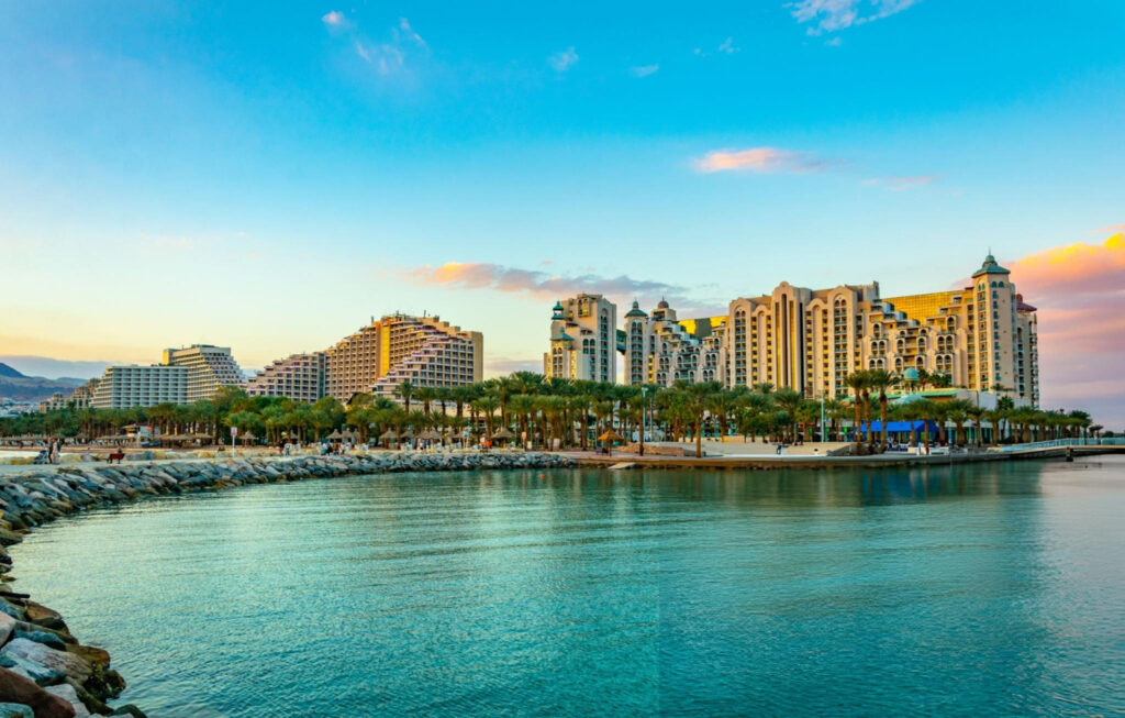 A scenic view of Eilat, a coastal city in Israel, featuring the turquoise waters of the Red Sea, sandy beaches, and a backdrop of modern buildings.