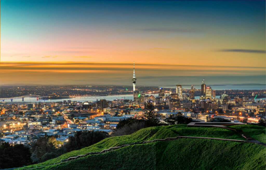 A panoramic view of Auckland City, New Zealand, featuring the skyline with the Sky Tower, modern skyscrapers, lush green parks, and Auckland Harbour