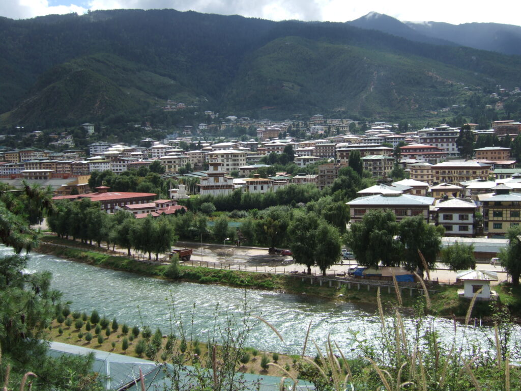 A panoramic view of Thimphu, the capital of Bhutan, featuring traditional Bhutanese architecture, lush green hills, and the Thimphu River flowing through the city