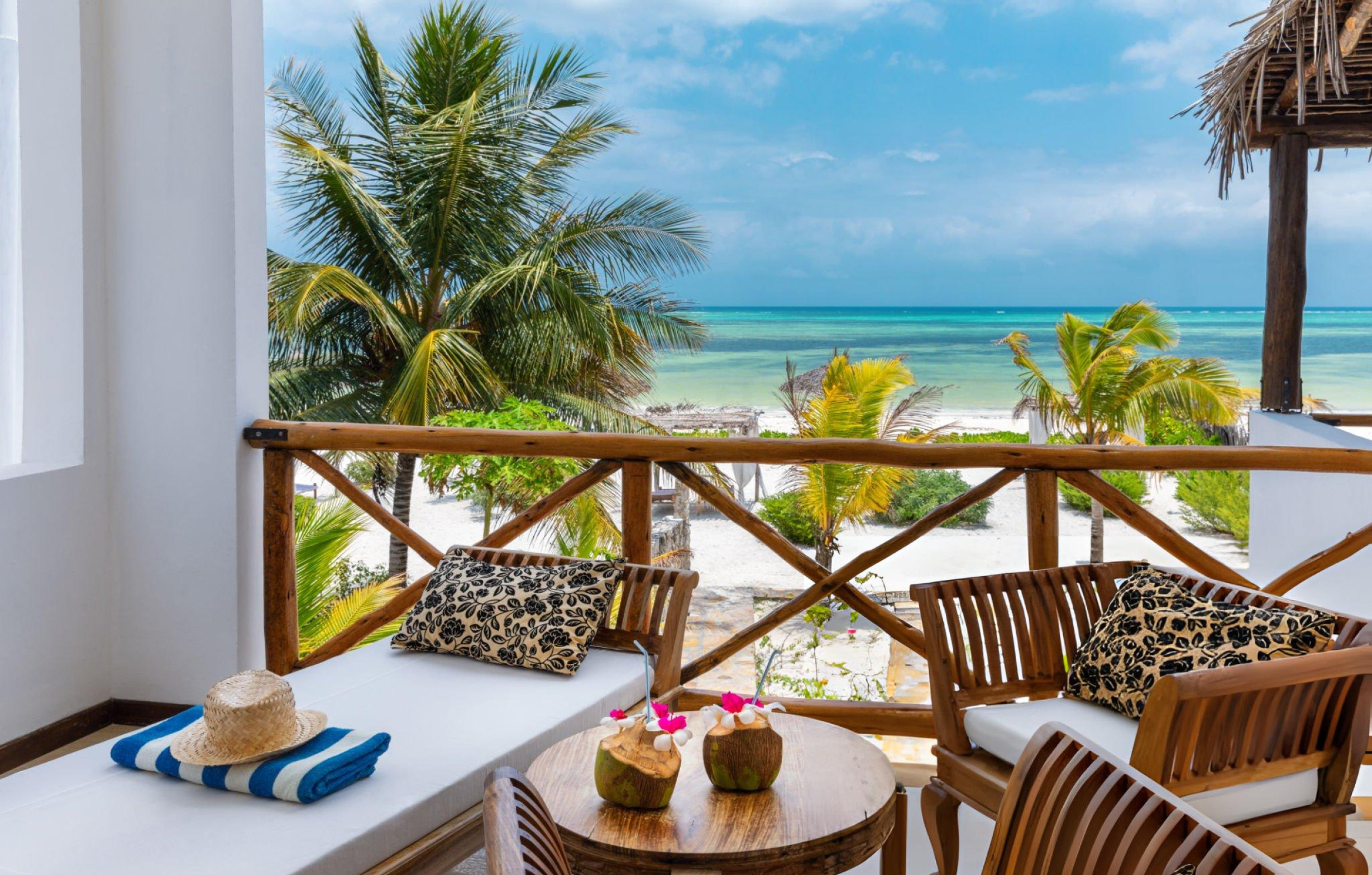 A serene view from a balcony overlooking Kendwa Beach, featuring palm trees, a sandy beach, and turquoise waters, with a cozy seating area adorned with cushions and fresh coconuts