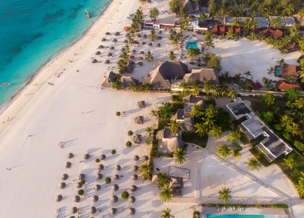 A picturesque view of Kendwa Beach, featuring soft white sands, clear turquoise waters, and palm trees lining the shoreline under a bright, sunny sky
