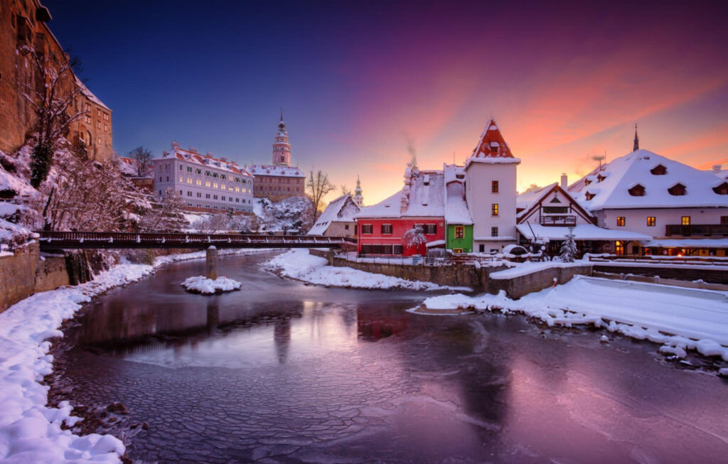 A scenic winter view of Český Krumlov, with snow-covered rooftops, the historic St. Vitus Church, and the Vltava River flowing through the town.