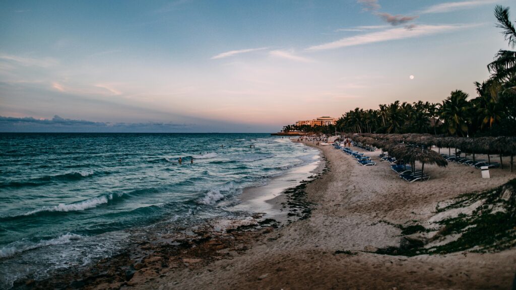 Wide view of a tranquil beach with white sand, clear blue water, and tropical palm trees under a bright sky.