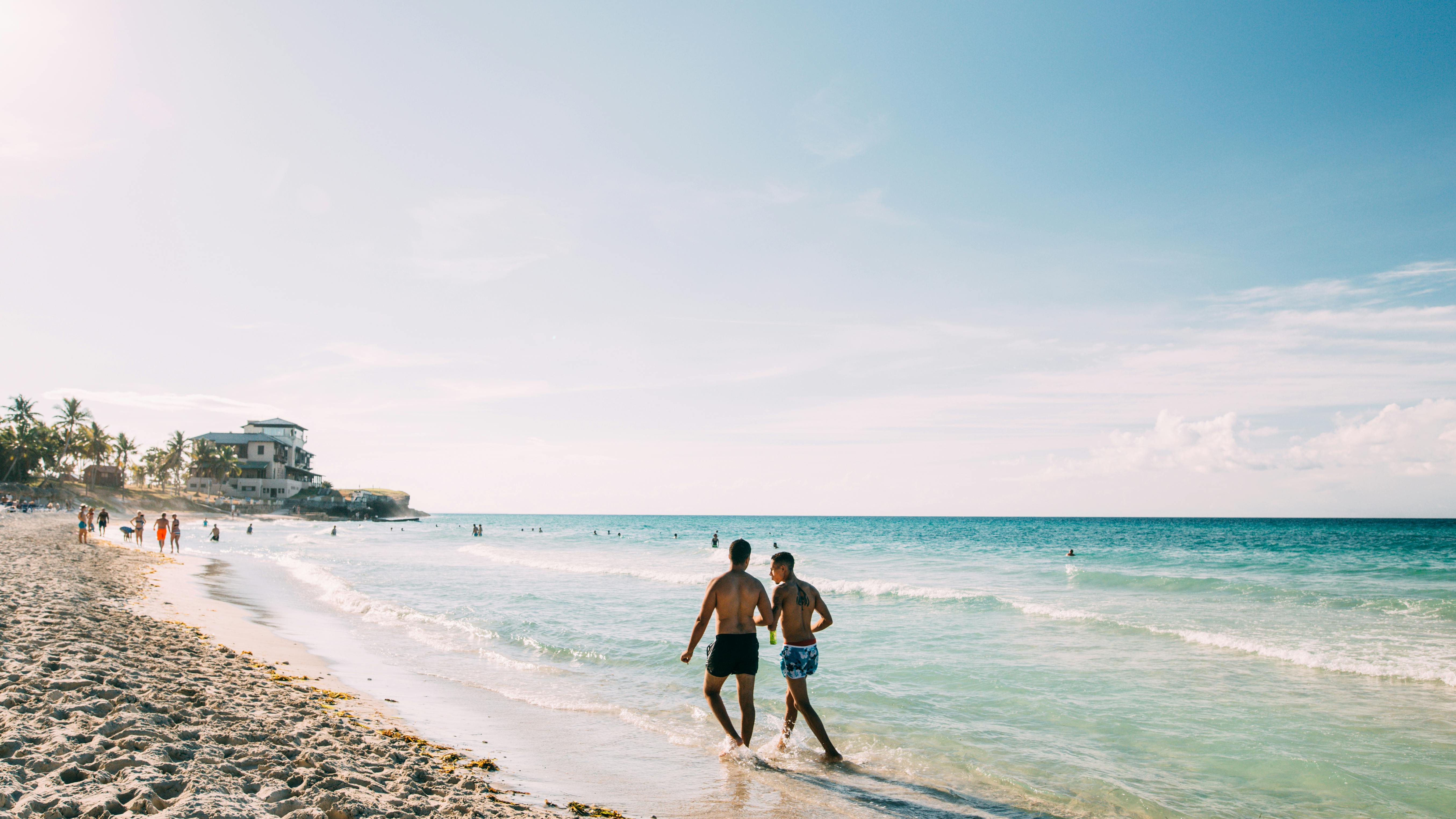 Sunlit view of Varadero Beach in Cuba with clear turquoise waters, white sandy shore, and lush green palm trees along the coastline.